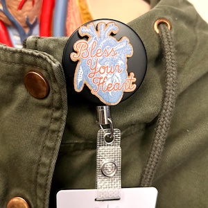 May include: A black retractable badge reel with a heart design and the words "Bless Your Heart" in a cursive font. The heart is blue with red accents. The reel is attached to a green jacket.