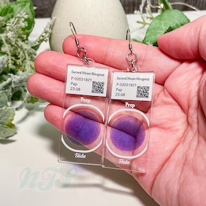 May include: A pair of clear acrylic earrings with a purple circle in the center. The earrings are shaped like microscope slides and have the text "Sacred Heart Hospital" and "Prep" printed on them.