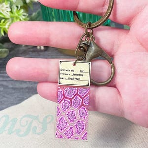 Op de afbeelding: Een roze en witte sleutelhanger met een vintage-stijl label waarop staat "Specimen No. 912, Locality: Queensland, Date: 16-03-1920". De sleutelhanger heeft een doorzichtig harsblok met een roze en wit patroon.