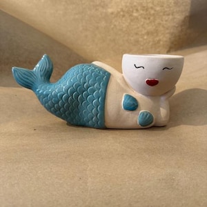 May include: A ceramic mermaid planter in a reclining pose. The mermaid has a blue tail with scale detailing, a white body, and a smiling face with red lips. Two blue seashell accents are on the chest. The planter is designed to hold small plants or succulents.