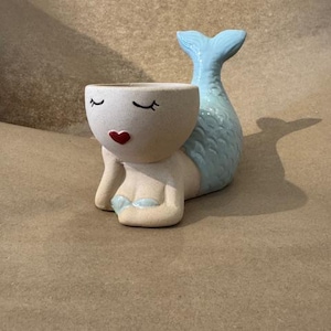 May include: A ceramic mermaid planter in a light beige color with a light blue tail. The mermaid has closed eyes, a red heart-shaped mouth, and light blue accents on the chest. The planter is designed to hold small plants or succulents.
