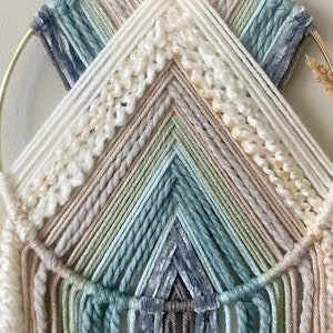 May include: A handcrafted wall hanging featuring a gold metal ring and a woven design. The piece incorporates various shades of white, beige, gray, and blue yarn, creating a textured, layered effect. The design includes a chevron pattern and fringe.