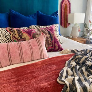 May include: A bed with a teal headboard and several pillows in shades of blue, pink, and patterned designs. A rust-colored throw blanket and a black and white striped blanket with pom-poms are at the foot of the bed. A macrame wall hanging and a lamp are also visible.