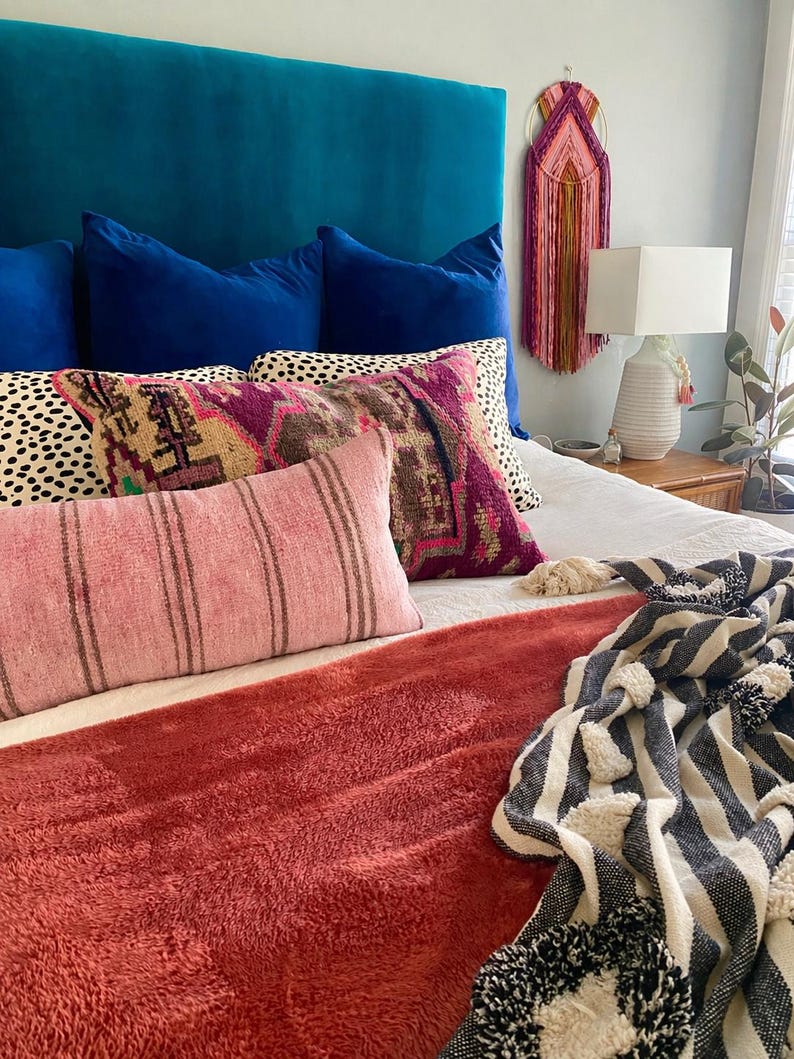 May include: A bed with a teal headboard and several pillows in shades of blue, pink, and patterned designs. A rust-colored throw blanket and a black and white striped blanket with pom-poms are at the foot of the bed. A macrame wall hanging and a lamp are also visible.