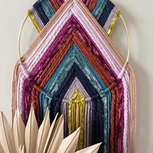 May include: A colorful macrame wall hanging with a gold hoop, featuring a geometric design with pink, red, orange, teal, blue, and yellow yarn. The hanging has long fringe strands.