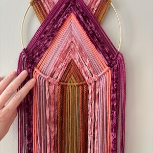May include: A handmade wall hanging featuring a gold metal ring and a colorful array of yarn in shades of pink, orange, and burgundy. The design incorporates geometric patterns and hanging strands, creating a textured, decorative piece.