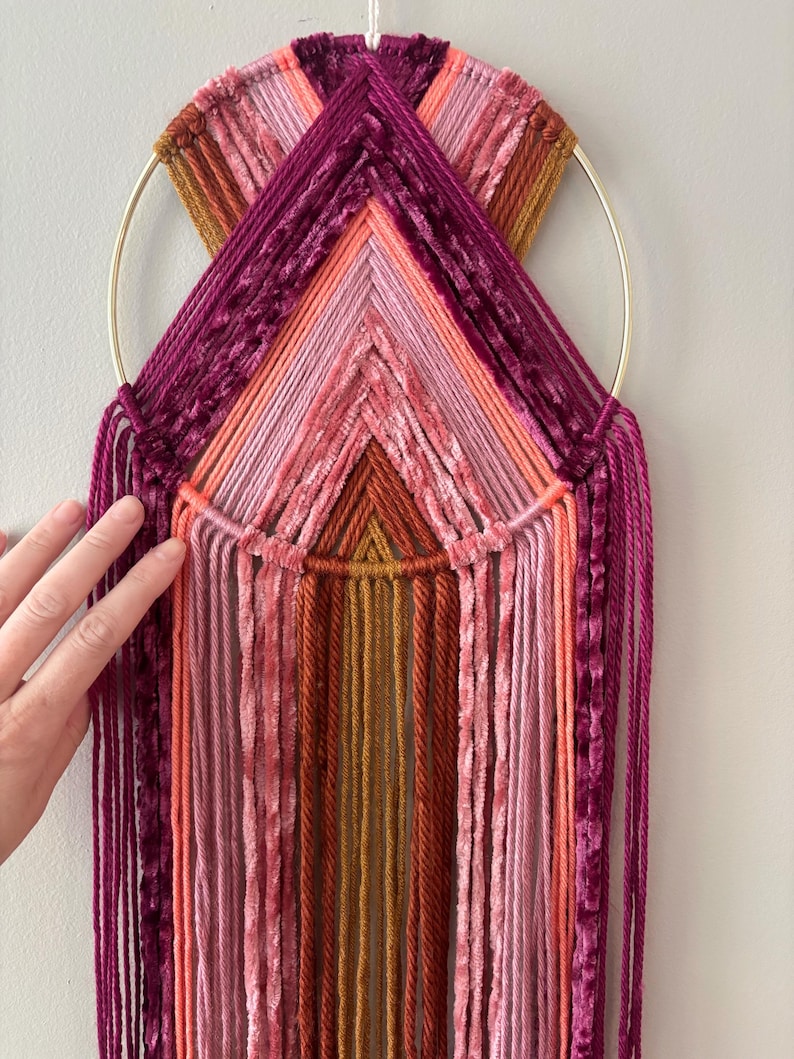 May include: A handmade wall hanging featuring a gold metal ring and a colorful array of yarn in shades of pink, orange, and burgundy. The design incorporates geometric patterns and hanging strands, creating a textured, decorative piece.