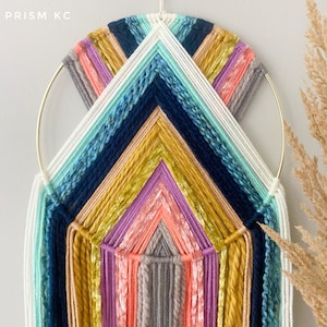 May include: A colorful macrame wall hanging with a geometric design. The hanging features a variety of colors, including blue, green, yellow, pink, and orange. The hanging is made with yarn and is attached to a gold hoop.
