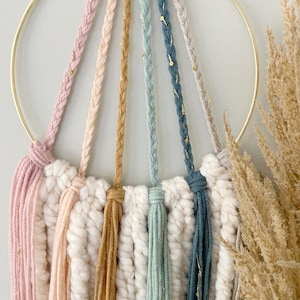 May include: A handcrafted macrame wall hanging featuring a gold metal ring and colorful yarn strands. The design includes pink, peach, tan, teal, and gray yarns, accented with gold thread. The bottom has a textured, off-white macrame base.