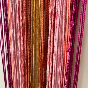 May include: Close-up of a wall hanging with vertical strands in shades of pink, orange, and gold. The textures vary from smooth to velvet, creating a visually interesting piece. The color palette is warm and inviting, suitable for home decor.