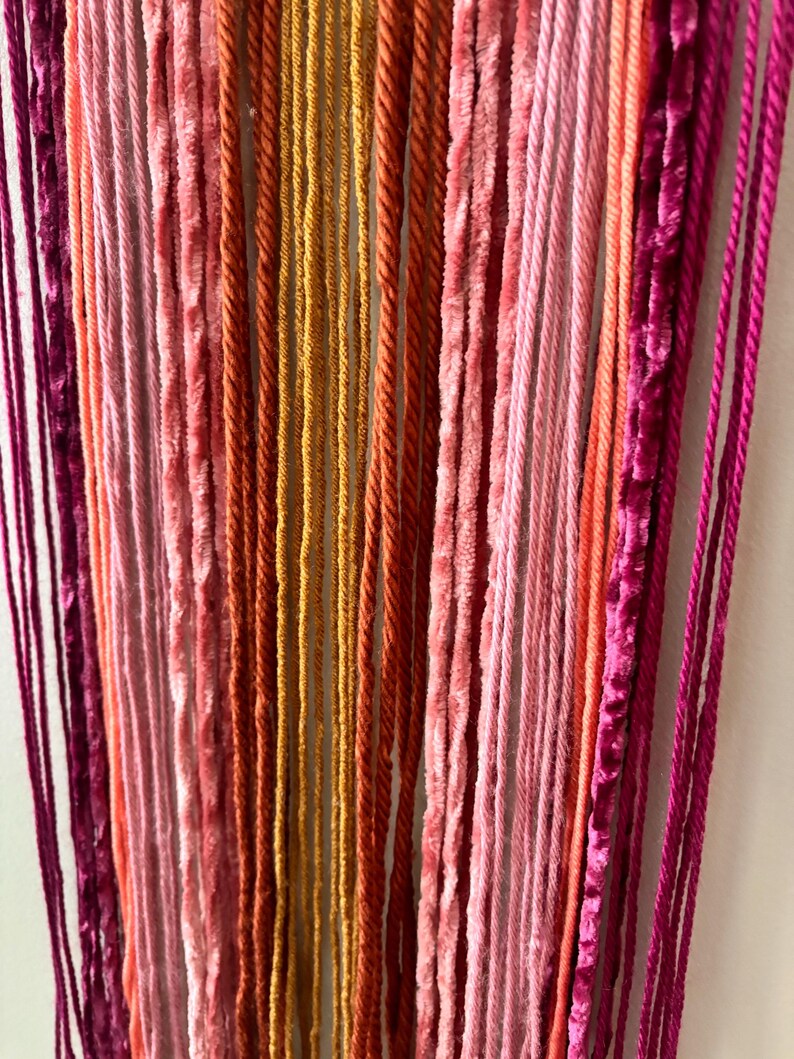 May include: Close-up of a wall hanging with vertical strands in shades of pink, orange, and gold. The textures vary from smooth to velvet, creating a visually interesting piece. The color palette is warm and inviting, suitable for home decor.