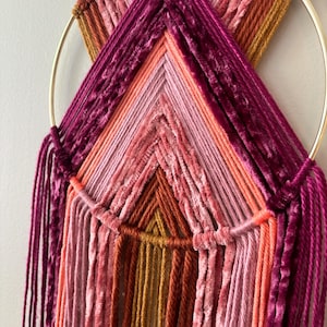 May include: A handmade wall hanging featuring a gold metal ring and a geometric design. The design incorporates various shades of pink, orange, and burgundy yarn, creating a textured and colorful piece of art. The yarn is arranged in a layered pattern.