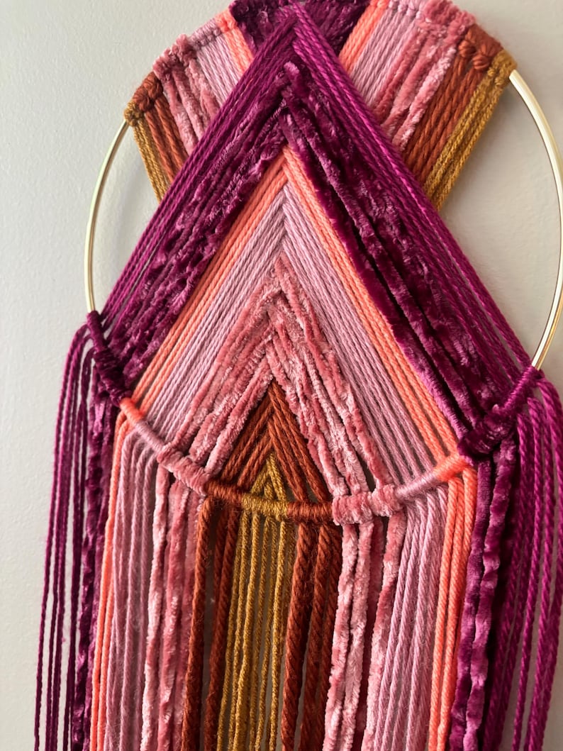 May include: A handmade wall hanging featuring a gold metal ring and a geometric design. The design incorporates various shades of pink, orange, and burgundy yarn, creating a textured and colorful piece of art. The yarn is arranged in a layered pattern.