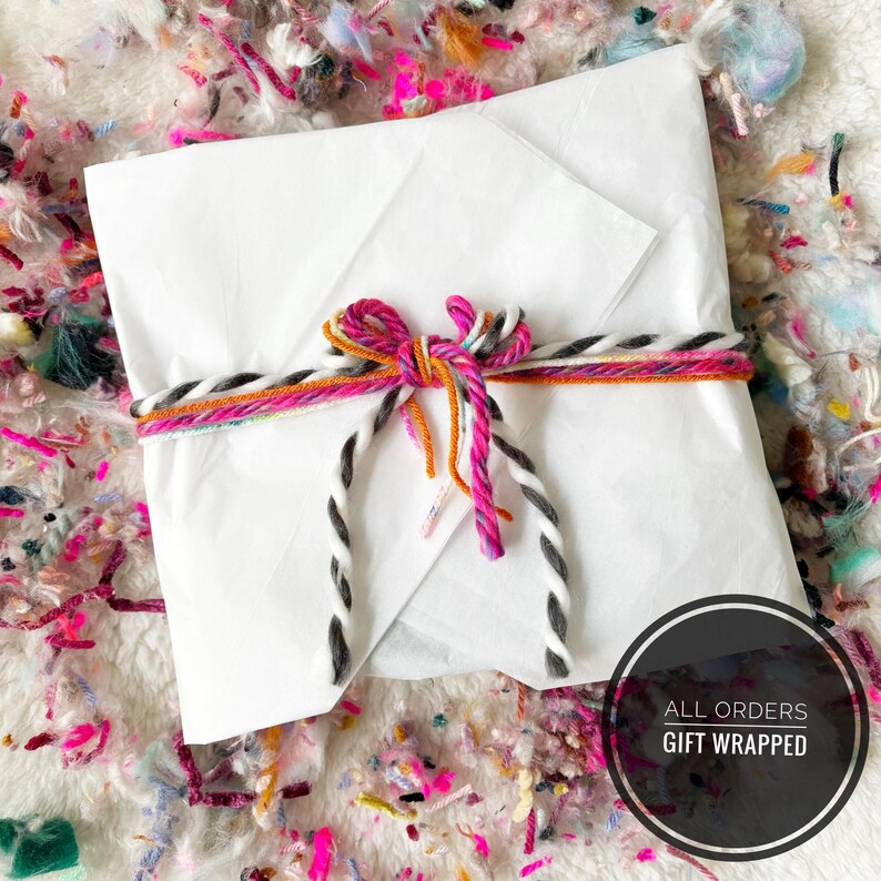 May include: A white package wrapped in white paper and tied with a colorful string bow. The bow features pink, orange, and gray accents. The package is labeled "ALL ORDERS GIFT WRAPPED" in a black circle.