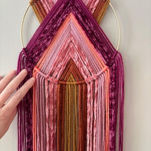May include: A handmade wall hanging featuring a gold metal ring and colorful yarn in shades of pink, orange, and burgundy. The piece measures 9" x 30" and has a geometric design with vertical fringe.