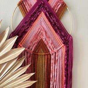 May include: A handcrafted wall hanging featuring a gold metal ring and a woven design. The piece incorporates various shades of pink, orange, and burgundy yarn, creating a textured and colorful geometric pattern. The design includes hanging strands.