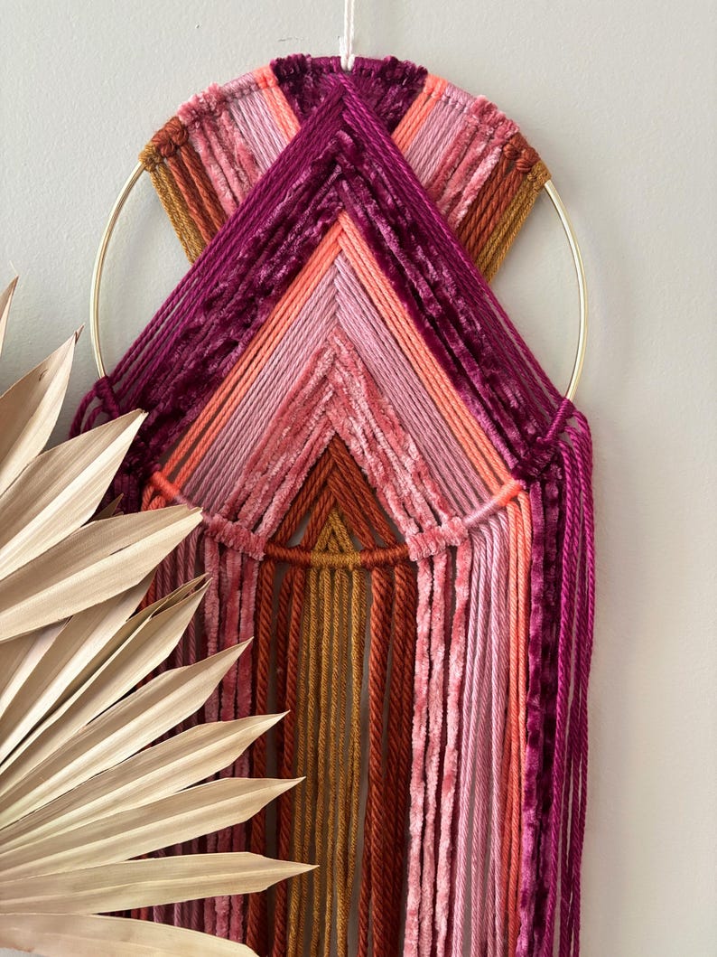 May include: A handcrafted wall hanging featuring a gold metal ring and a woven design. The piece incorporates various shades of pink, orange, and burgundy yarn, creating a textured and colorful geometric pattern. The design includes hanging strands.