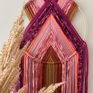 May include: A handmade wall hanging featuring a gold metal ring and a geometric design. The piece uses various shades of pink, orange, and burgundy yarn and velvet, creating a textured and colorful artwork. Decorative pampas grass is in the foreground.