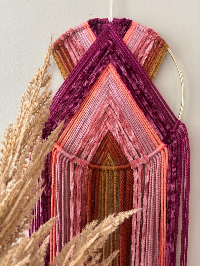 May include: A handmade wall hanging featuring a gold metal ring and a geometric design. The piece uses various shades of pink, orange, and burgundy yarn and velvet, creating a textured and colorful artwork. Decorative pampas grass is in the foreground.