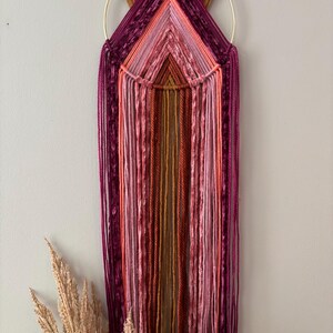 May include: A handmade wall hanging featuring a gold metal ring and a geometric design. The artwork uses various shades of pink, purple, and orange yarn, creating a textured and colorful piece. The yarn strands hang vertically, adding a bohemian touch.