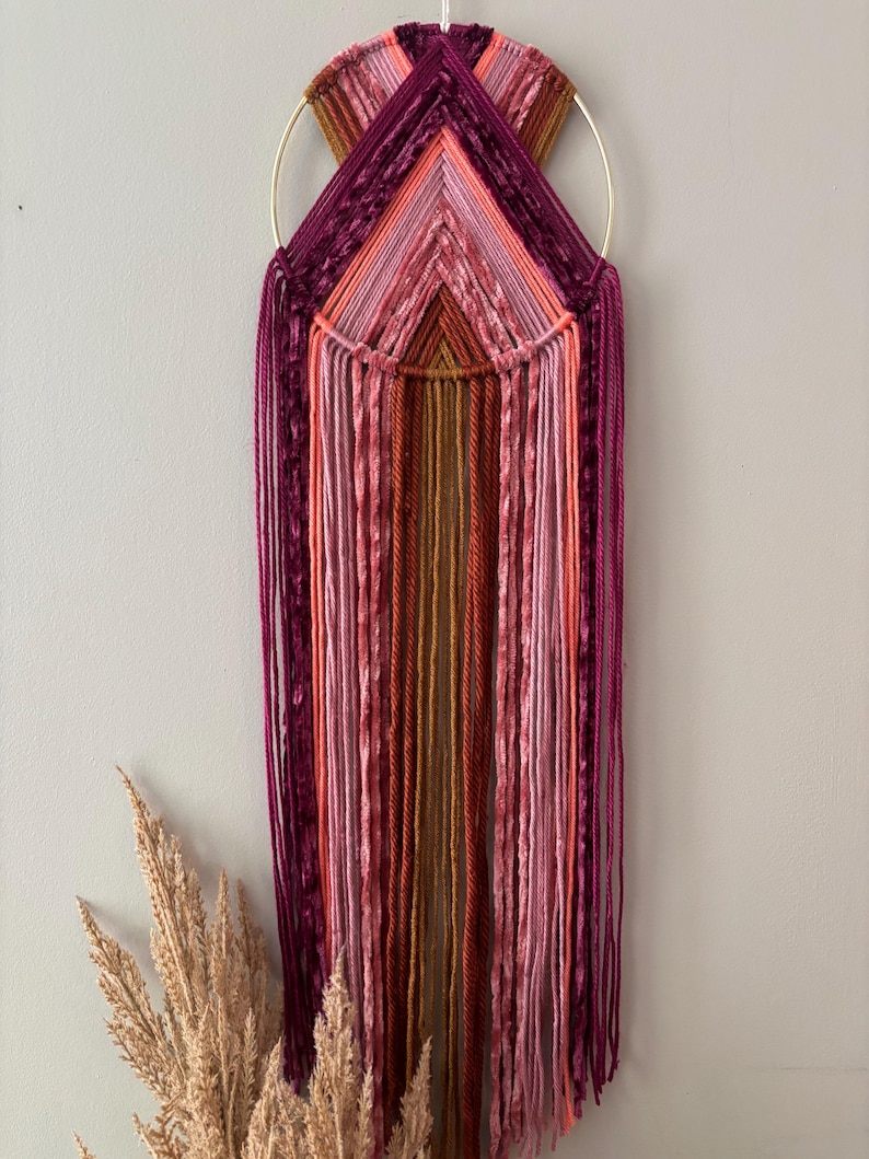 May include: A handmade wall hanging featuring a gold metal ring and a geometric design. The artwork uses various shades of pink, purple, and orange yarn, creating a textured and colorful piece. The yarn strands hang vertically, adding a bohemian touch.