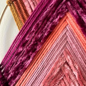 May include: A close-up of a handmade wall hanging with a circular metal frame. The artwork features a vibrant array of colors, including shades of pink, orange, and burgundy, woven in a geometric pattern. The texture is a mix of smooth and plush yarns.