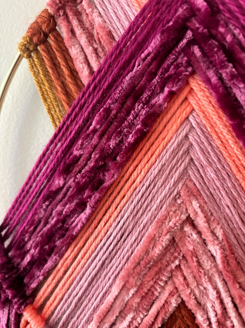 May include: A close-up of a handmade wall hanging with a circular metal frame. The artwork features a vibrant array of colors, including shades of pink, orange, and burgundy, woven in a geometric pattern. The texture is a mix of smooth and plush yarns.