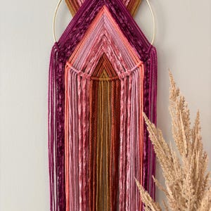 May include: A handcrafted wall hanging featuring a gold metal ring and a geometric design. The piece uses various shades of pink, orange, and burgundy yarn, creating a textured, layered effect. Long strands of yarn cascade downwards, adding a bohemian touch to the decor.