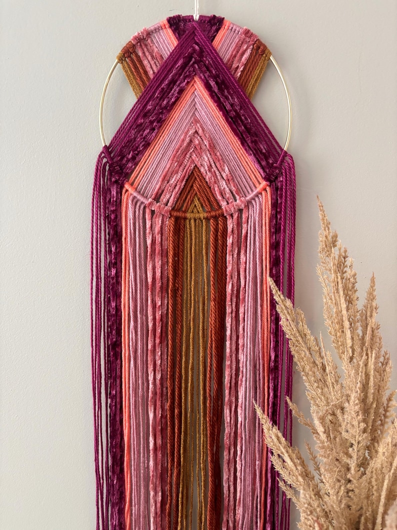 May include: A handcrafted wall hanging featuring a gold metal ring and a geometric design. The piece uses various shades of pink, orange, and burgundy yarn, creating a textured, layered effect. Long strands of yarn cascade downwards, adding a bohemian touch to the decor.