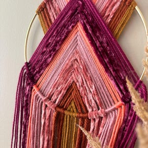 May include: A handmade wall hanging featuring a gold metal ring and a woven design. The piece uses various shades of pink, orange, and burgundy yarn, creating a textured, geometric pattern. Strands of yarn hang from the bottom, adding to the bohemian aesthetic.