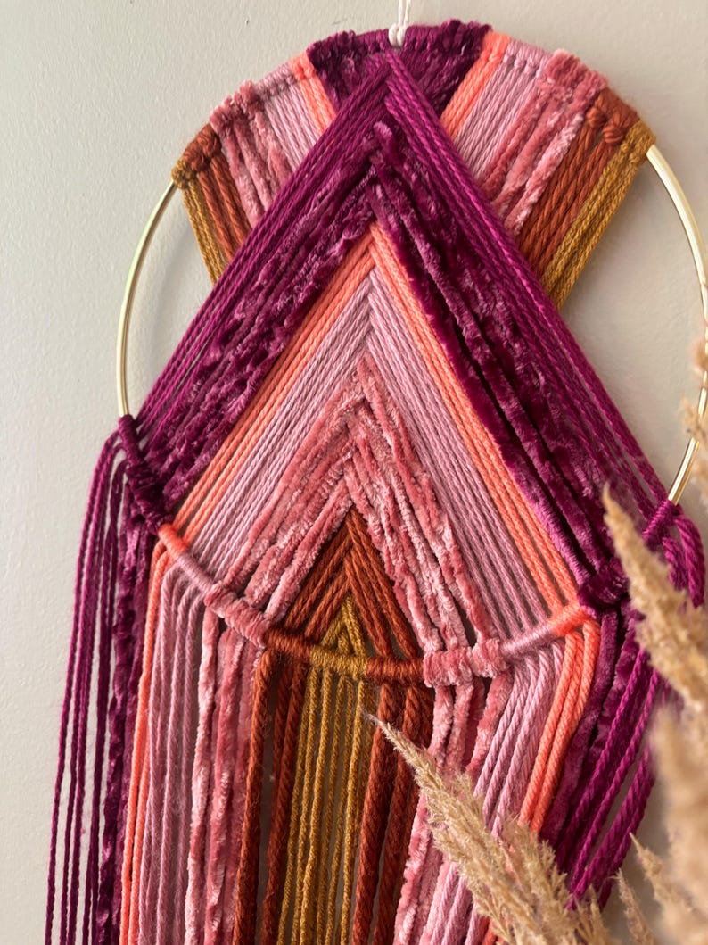 May include: A handmade wall hanging featuring a gold metal ring and a woven design. The piece uses various shades of pink, orange, and burgundy yarn, creating a textured, geometric pattern. Strands of yarn hang from the bottom, adding to the bohemian aesthetic.