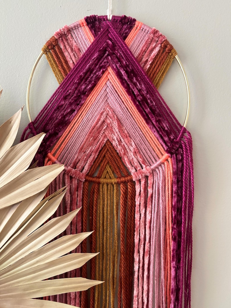 May include: A handmade wall hanging featuring a gold metal ring and a woven design in shades of pink, orange, and burgundy. The textured yarn creates a geometric pattern, adding a touch of bohemian flair to any space.