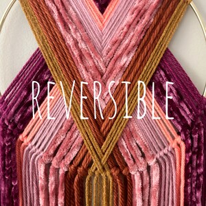 May include: A handmade wall hanging featuring a gold metal ring and a woven design. The piece showcases a gradient of colors, including shades of pink, orange, and burgundy. The word "REVERSIBLE" is displayed in white text.
