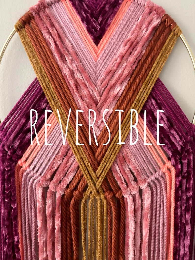 May include: A handmade wall hanging featuring a gold metal ring and a woven design. The piece showcases a gradient of colors, including shades of pink, orange, and burgundy. The word "REVERSIBLE" is displayed in white text.