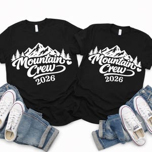 Puede incluir: Dos camisetas negras con el texto blanco "Mountain Crew 2026", con un dise&ntilde;o de monta&ntilde;a y &aacute;rboles. Las camisetas se combinan con vaqueros azules y zapatillas de lona blancas.