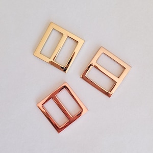 May include: Three metal belt buckles in gold, rose gold, and copper colours. Each buckle has a square shape with two rectangular openings. The buckles are arranged on a white surface.