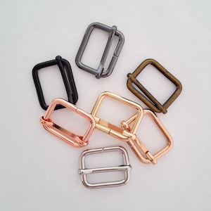 May include: A selection of rectangular metal buckles in black, gunmetal, bronze, rose gold, gold, and silver finishes. These are used for bags, belts, and crafts. The buckles are displayed on a white background.