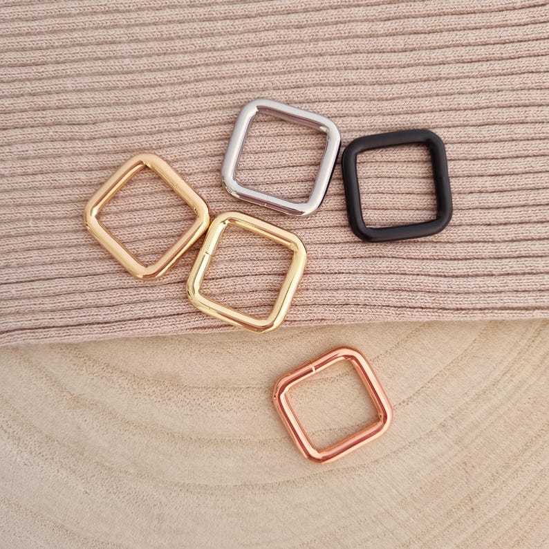 Peut inclure: Plusieurs anneaux carr&eacute;s en m&eacute;tal de diff&eacute;rentes couleurs, dont or, argent, noir et or rose. Les anneaux sont dispos&eacute;s sur un tissu de couleur neutre, mettant en valeur leurs diff&eacute;rentes finitions et formes. Ces anneaux sont probablement utilis&eacute;s pour l'artisanat ou les accessoires.