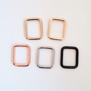 May include: Five rectangular metal buckles in various finishes: rose gold, gold, silver, and black. The buckles are arranged on a white surface. These hardware components are likely used in bag making or other crafting projects.