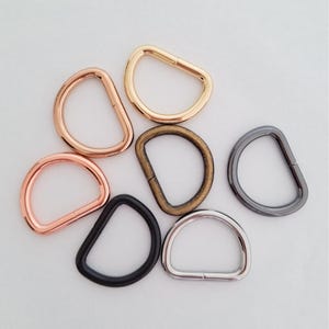 May include: A collection of seven D-rings in various metallic finishes, including rose gold, polished gold, antique brass, matte black, gunmetal, and silver. Each metal D-ring features a smooth, rounded shape with a visible seam on the straight edge.