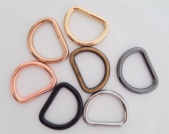 May include: A collection of seven D-rings in various metallic finishes, including rose gold, polished gold, antique brass, matte black, gunmetal, and silver. Each metal D-ring features a smooth, rounded shape with a visible seam on the straight edge.