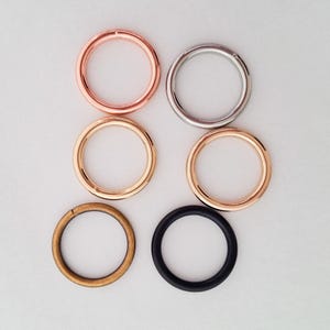 May include: Six metal rings in various colors: rose gold, silver, gold, and black. The rings are arranged in two rows of three, showcasing different finishes and materials. Ideal for crafting or jewelry making.