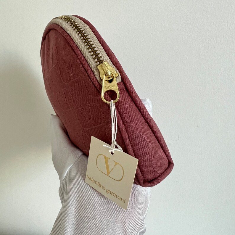 Peut inclure: Une petite trousse de maquillage ronde de couleur bordeaux avec une fermeture &eacute;clair dor&eacute;e et une &eacute;tiquette blanche sur laquelle est inscrit "valentino garavani".