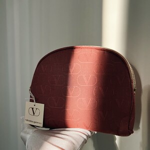 Peut inclure: Une trousse de maquillage rouge avec une fermeture &eacute;clair dor&eacute;e et un motif "V" r&eacute;p&eacute;t&eacute;. La trousse est munie d'une &eacute;tiquette blanche avec le texte "valentino garavani" et un grand logo "V".