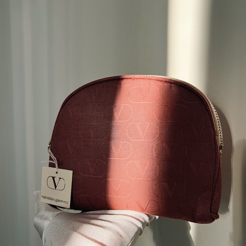 Peut inclure: Une trousse de maquillage rouge avec une fermeture &eacute;clair dor&eacute;e et un motif "V" r&eacute;p&eacute;t&eacute;. La trousse est munie d'une &eacute;tiquette blanche avec le texte "valentino garavani" et un grand logo "V".