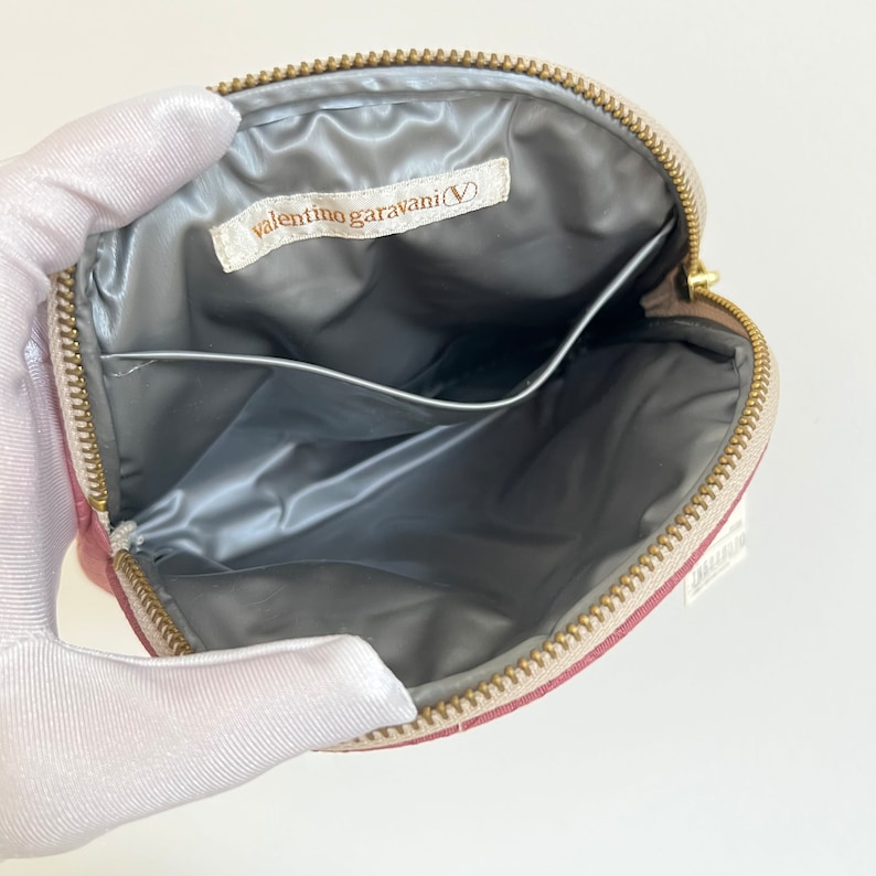 Peut inclure: L'int&eacute;rieur d'une trousse de maquillage ronde rose avec une fermeture &agrave; glissi&egrave;re. La trousse est doubl&eacute;e de satin gris et poss&egrave;de une petite poche. La trousse porte une &eacute;tiquette indiquant "valentino garavani(V)"