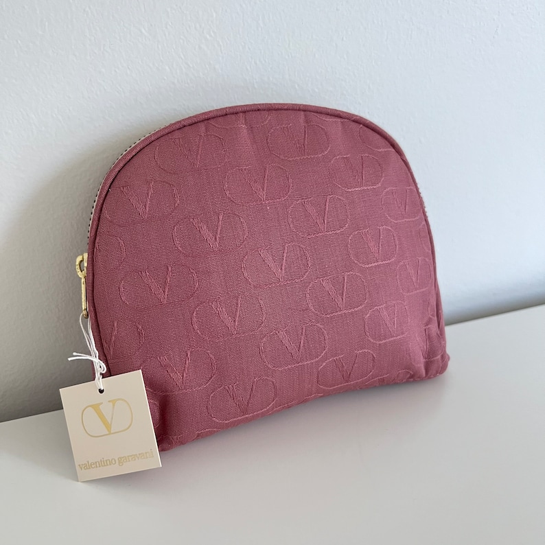 Peut inclure: Une trousse de maquillage rose avec un motif en relief r&eacute;p&eacute;t&eacute; en forme de "V". La trousse est dot&eacute;e d'une fermeture &eacute;clair dor&eacute;e et d'une &eacute;tiquette blanche avec le texte "valentino garavani".