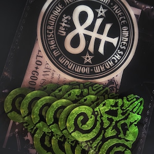 May include: A close-up of a sticker with a black and white symbol of an infinity symbol with a cross through it. The sticker has the text "MAGISTRUMQUE ATRUM MEUM AVECE OMNES SACANAM-DOMINUM" around the symbol.