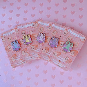 May include: Five enamel pins featuring a cartoon frog design in various colors: blue, yellow, purple, pink, and green. Each frog wears a dress and has large eyes. The pins are displayed on pink backing cards with a bread and heart pattern.