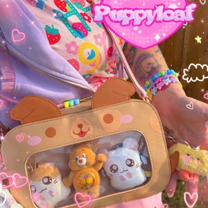 May include: Tan dog-shaped crossbody bag with a clear window displaying three plush toys. The toys include a hamster, a bear, and a white bunny. A yellow plush keychain hangs from the bag. The bag has a pink strap.